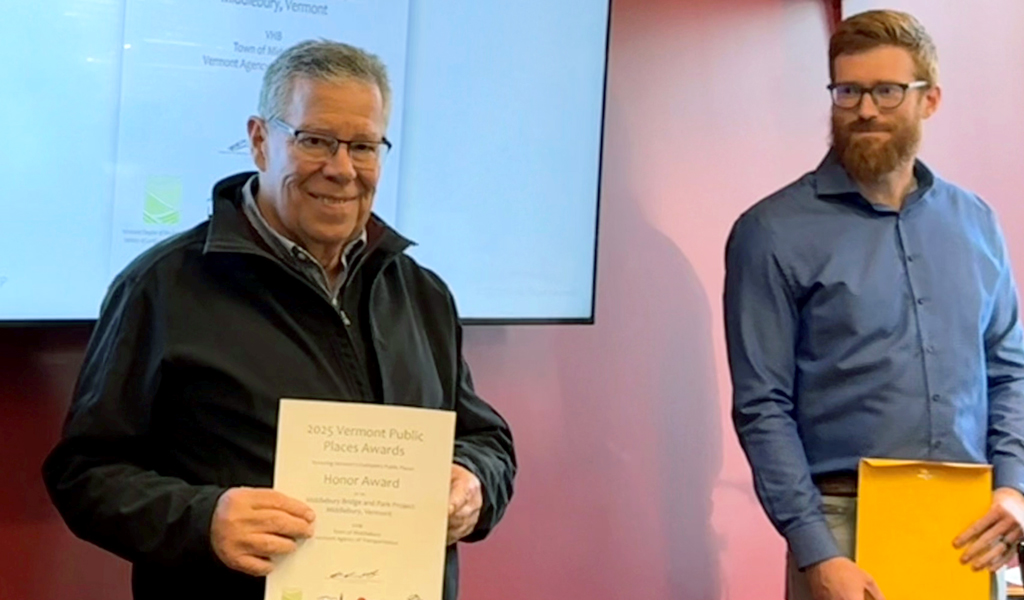 Two individuals are smiling and standing in front of a presentation screen displaying the "2025 Vermont Public Places Awards."