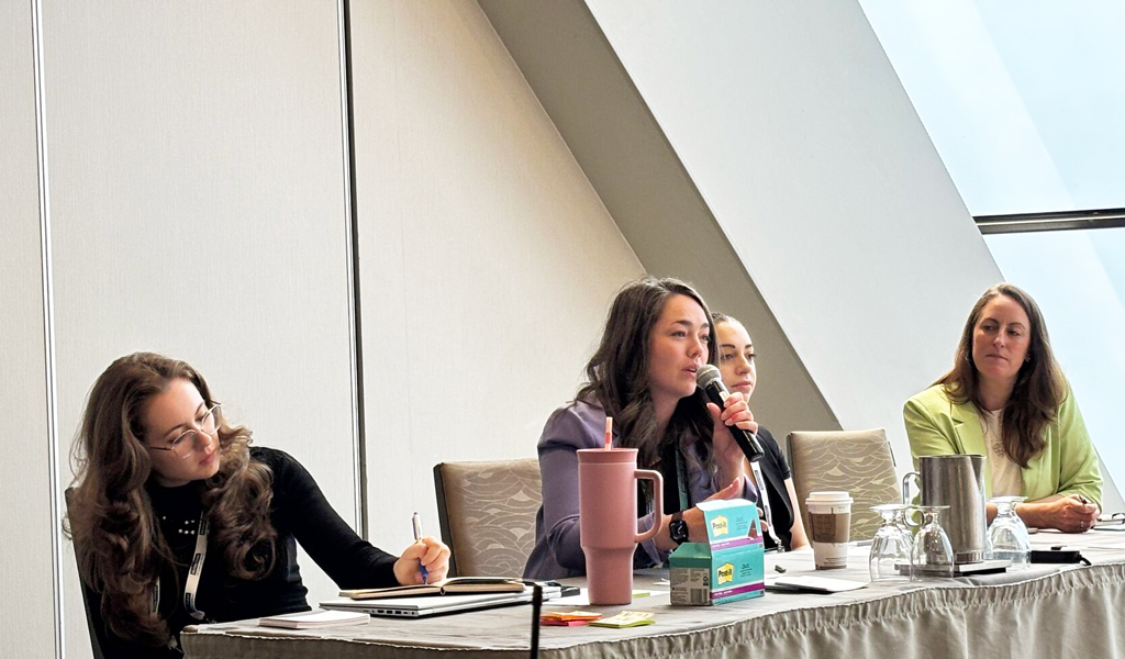 Four individuals participate in a panel discussion at a conference room table, equipped with microphones and laptops.