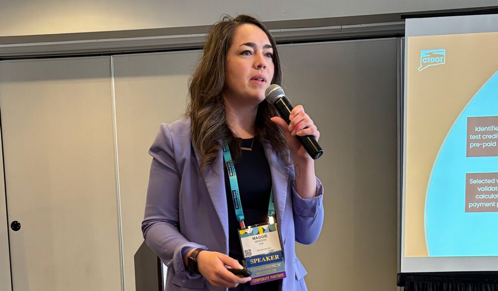 A person presenting at a conference, standing with a microphone in front of a projected presentation slide.