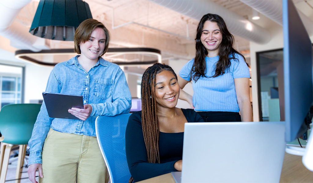 Three employees, standing and sitting, gathered around a computer.