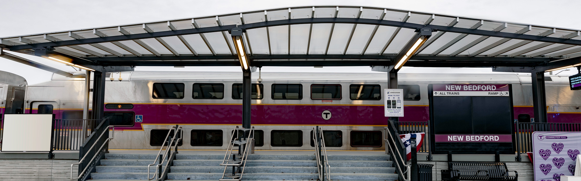 A train station covered by a modern canopy, with an MBTA commuter train in the background.